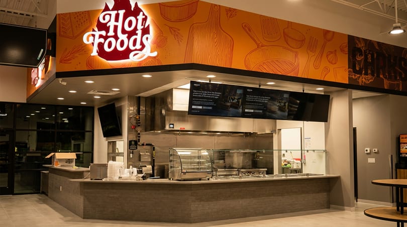 Bright 'Hot Foods' counter with a flame logo sign, digital menus, and stainless steel kitchen equipment.