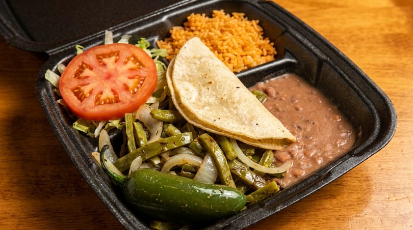 Black takeout container with a taco, rice, refried beans, nopales, tomato, lettuce, and a jalapeño.