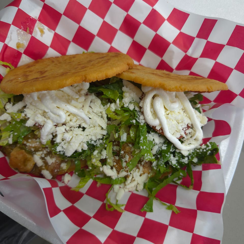 A gordita with lettuce, cheese, and cream, served in a red and white checkered paper.