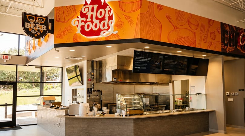 Modern restaurant interior with "Hot Foods" and "Draft Beer" signs, kitchen counter, and orange cooking mural.