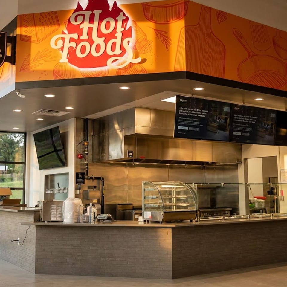 Restaurant interior with "Hot Foods" and "Draft Beer Here" signs, a kitchen counter, and large windows.