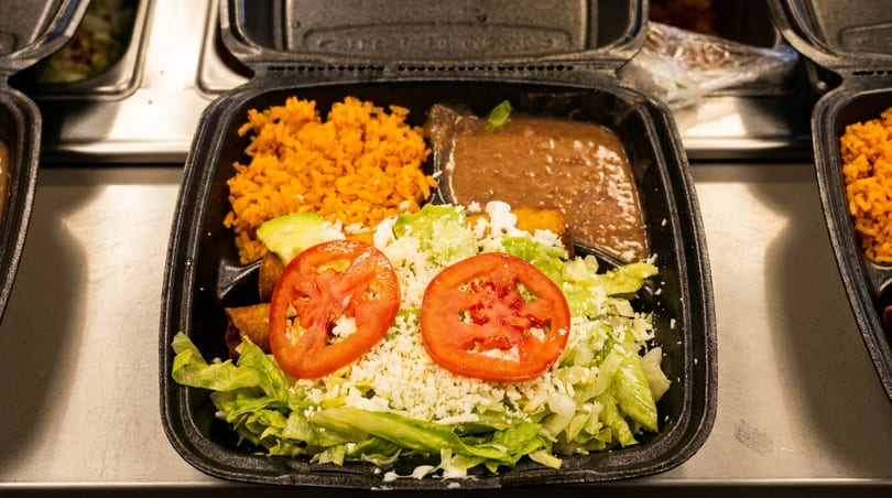 Mexican takeout meal with orange rice, refried beans, salad, and fried taquitos in a black container.