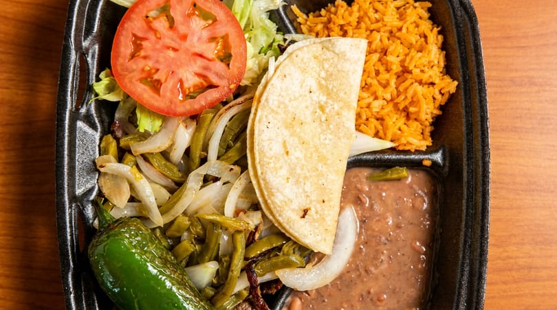Mexican meal in a takeout container with corn tortilla, rice, beans, sautéed vegetables, tomato, and chili.
